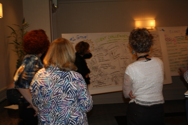 Viele Frauen stehen vor einer weißen Tafel mit Text, unter einer Wandlampe, mit einer Pflanze links, in einem Raum.