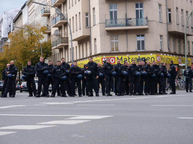 Eine Gruppe von Polizisten in schwarzen Uniformen mit blauen Helmen steht in einer Straße, die von Laternenmasten, Bäumen und Glasfenstergebäuden gesäumt ist, unter einem klaren blauen Himmel.