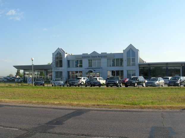 Ein Autohaus mit parkenden Autos davor, umgeben von Gebäuden, Straßeninfrastruktur, fahrenden Fahrzeugen und Bäumen unter einem bewölkten Himmel.