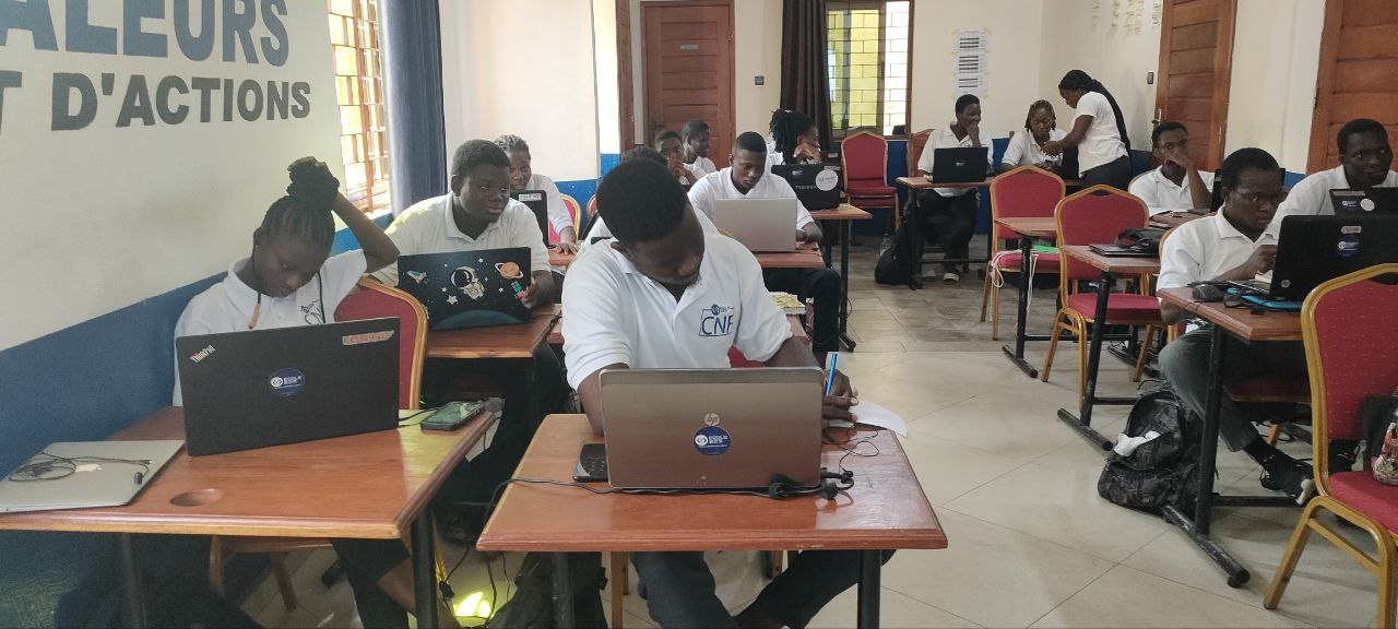 Eine Gruppe von Schülern sitzt an Tischen mit Laptops in einem Klassenzimmer, mit Taschen auf dem Boden und einer Wand mit Text auf der linken Seite, Fenstern und Türen im Hintergrund, was auf eine Online-Kurs-Situation in einer Schule in Ghana hinweist.