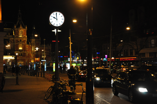 Eine nächtliche StraßenSzene mit Fahrzeugen auf der Straße, Fahrrädern neben Pfählen, einer Uhr an einem Pfahl, beleuchteten Gebäuden im Hintergrund und Menschen auf dem linken Gehweg.
