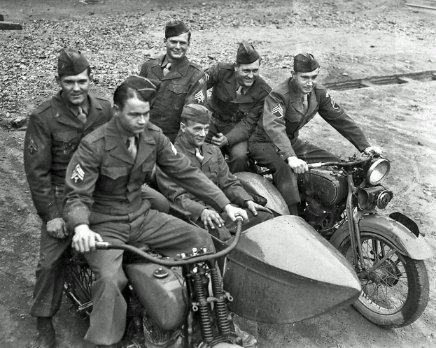 Ein Schwarz-Weiß-Foto von einer Gruppe von Männern, die auf Fahrrädern sitzen und lächeln, auf einer Straße.