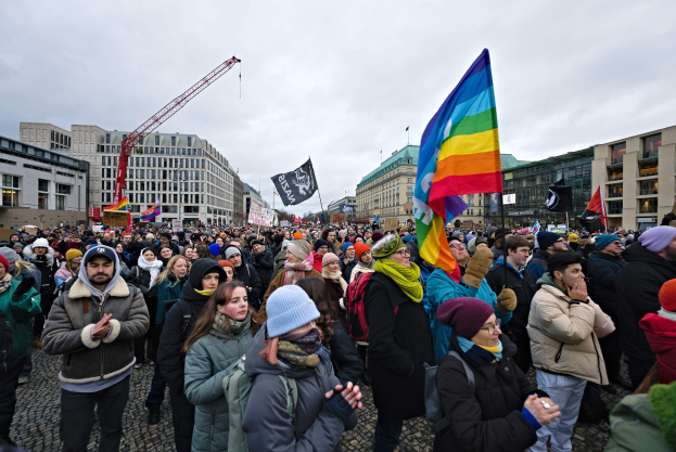 Große Gruppe von Menschen bei einer LGBTQ+-Rechtsdemo in Berlin, die Fahnen und Schilder schwingt, mit Gebäuden, einem Kran und Wolken im Hintergrund.