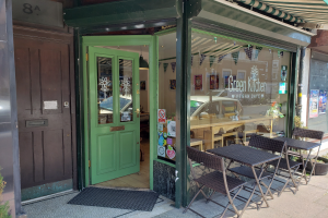 Ein Außenbereich mit Tischen und Stühlen, umgeben von Pflanzen, einer Fußmatte und einer Glaswand mit Postern, der in das Green Kitchen Vegan Cafe mit Innen-Tischen, -Stühlen und einer Deckenlampe führt.