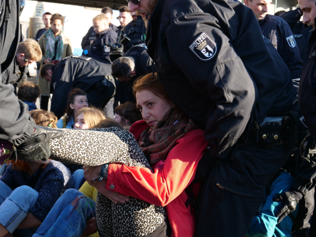 Eine Frau in dunkler Kleidung wird von einem Polizeibeamten in Uniform begleitet, hinter ihnen sitzen und stehen Menschen, im Hintergrund ein Schuppen und ein klarer blauer Himmel.