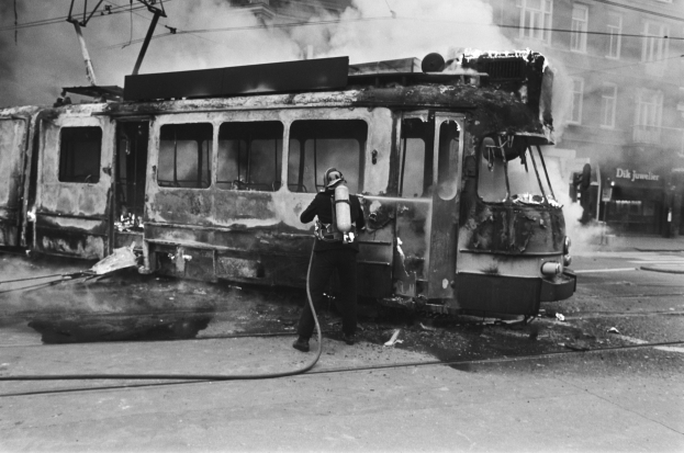 Schwar├č-we├čüsses Bild eines Mannes in einem Helm, der eine Pfeife h├Ąlt und neben einer ausgebrannten Stra├čenbahn auf einer Stra├če mit aufsteigendem Rauch, ├ťberkopfdr├Ąhten und einem Geb├Ąude im Hintergrund steht.