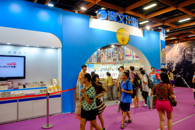 Eine Gruppe von Menschen steht vor einem Messestand mit einem Fernseher links, einer Absperrung und Hintergrundplakaten mit Text, Bildern und Deckenlampen.