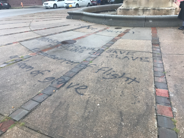 Geweg mit 'Black Lives Matter'-Graffiti in großen Buchstaben vor einem Gebäude, mit Fahrzeugen, Menschen, Bäumen, Pfählen und einer Wand in der Nähe.