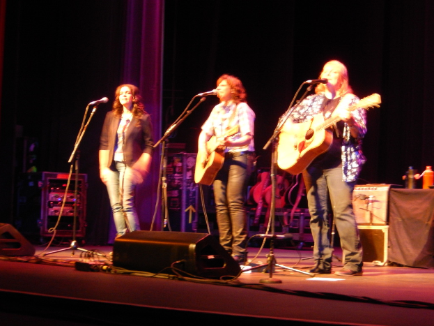 Drei Frauen performen auf der Bühne bei einem Musikkonzert, jede singt in ein Mikrofon, während sie eine Gitarre spielen, mit einem Tisch mit Wasserflaschen und zusätzlichen Gitarren dahinter.