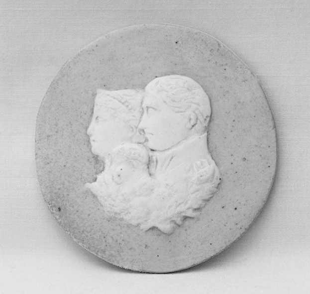 Ein Schwarz-Weiß-Foto eines Mannes in einem Anzug, der ein Baby hält, mit einer Frau im Profil neben ihm, alles mit unscharfem Hintergrund.