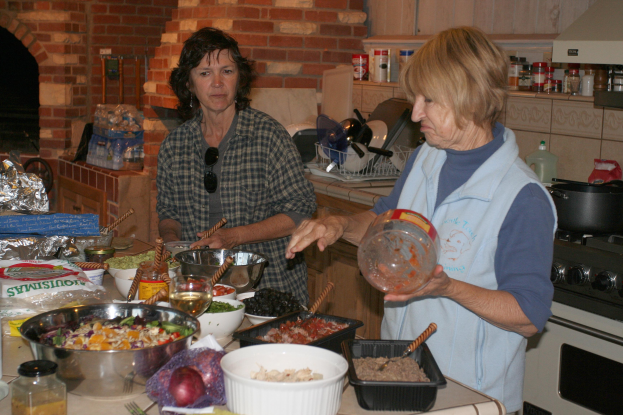 Zwei Frauen stehen in einer Küche umgeben von verschiedenen Lebensmitteln und Küchenutensilien auf der Arbeitsplatte, mit einem Gasherd auf der rechten Seite und Flaschen, einem Kamin und einer Backsteinwand im Hintergrund, bereiten gemeinsam ein Essen zu.