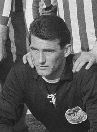 Schwarzes und weißes Foto eines Mannes, der auf einem grünen Fussballfeld sitzend eine T-Shirt und eine Kette trägt, mit zwei Personen hinter ihm.