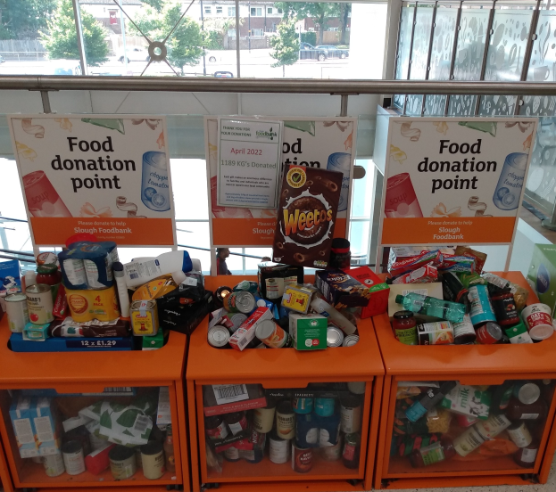 Orange Schränke mit gespendeten Lebensmitteln wie Flaschen, Dosen und Kartons, mit Informationsschildern, durch Glasfenster mit Fahrzeugen, Bäumen, Gebäuden und Himmel.