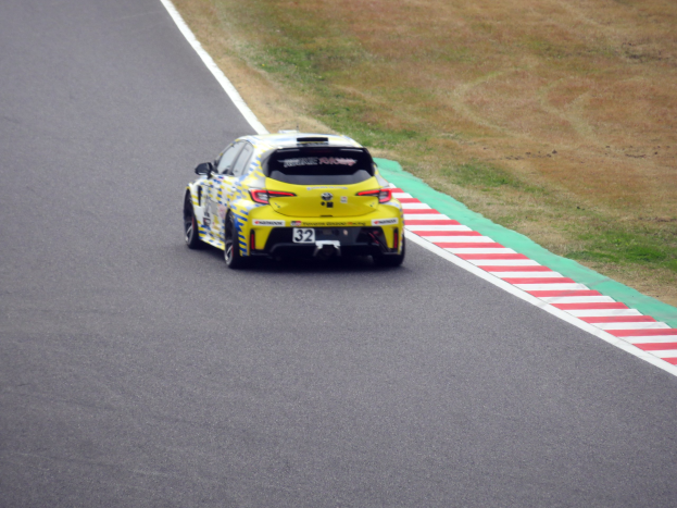 Ein gelber Sportwagen mit Text und Zahlen darauf, der eine Rennstrecke entlangfährt, umgeben von Gras.