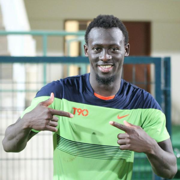 Ein Mann in einem grün-blauen Hemd, identifiziert als Emmanuel Adebayor, zeigt mit einem Lächeln auf die Kamera, mit einem unscharfen Geländer, einer Wand und einer Tür im Hintergrund.