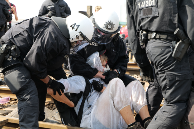 Eine Gruppe von Polizisten in Einsatzausrüstung verhaftet einen Mann in einem weißen Kleid auf einem Bahngleis, mit anderen Menschen im Hintergrund.