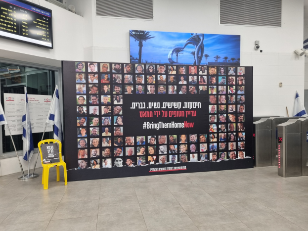 Eine große Wand zeigt zahlreiche Fotos, einen gelben Stuhl, israelische Regierungsflaggen auf Stangen, eine Tafel mit dem Text 'Bring Them Home Now', einen montierten Bildschirm, Deckenleuchten und andere Gegenstände.