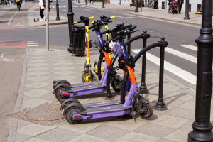 Eine Reihe von Elektro-Scootern, die entlang einer Straße mit Fußgängern, Radfahrern, einem Müllcontainer und im Hintergrund Gebäuden, Bäumen und Pflanzen geparkt sind.