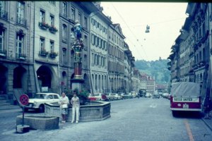 Alte Schwarz-Weiß-Fotografie einer Stadtstraße mit Fußgängern, geparkten Autos, Gebäuden, einer Statue auf einem Sockel, einem Wegweiser, Topfpflanzen, Bäumen und einem klaren blauen Himmel.