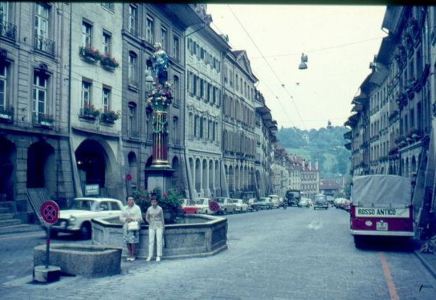 Alte Schwarz-Weiß-Fotografie einer Stadtstraße mit Fußgängern, geparkten Autos, Gebäuden, einer Statue auf einem Sockel, einem Wegweiser, Topfpflanzen, Bäumen und einem klaren blauen Himmel.
