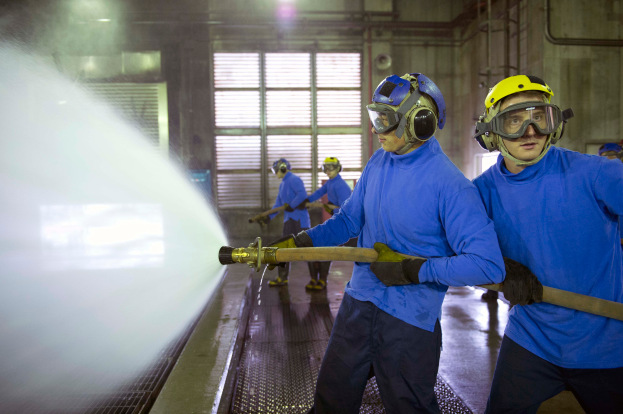 Eine Gruppe von Männern in blauen Hemden und gelben Helmen arbeitet an einer Maschine in einer Fabrikumgebung, wobei einer der Männer mit einem Rohr Wasser auf den Boden sprüht.