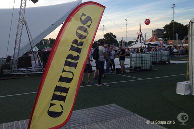 Ein buntes Karnevalsszenario mit Zelten, einem zentralen Mast, Menschen, Bäumen, Strommasten, Straßenbahnen auf der Straße und einem Eisenständer auf der linken Seite unter einem klaren Himmel.