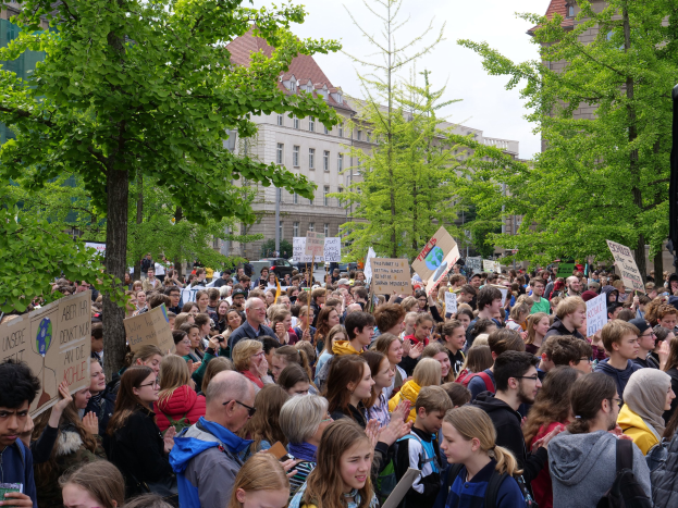 Große Menschenmenge protestiert vor einem Gebäude in Berlin, hält Schilder, mit Bäumen, Fahrzeugen, einem Lautsprecher und Himmel