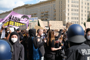Eine Gruppe maskierter Demonstranten mit Schildern vor einem Gebäude, mit zwei helmträgern Polizisten rechts daneben, vor einem Hintergrund aus Bäumen und einer bewölkten Himmel.