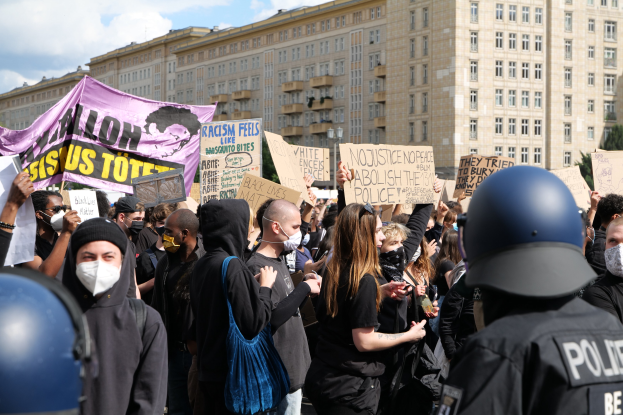 Eine Gruppe maskierter Demonstranten mit Schildern vor einem Gebäude, mit zwei helmträgern Polizisten rechts daneben, vor einem Hintergrund aus Bäumen und einer bewölkten Himmel.
