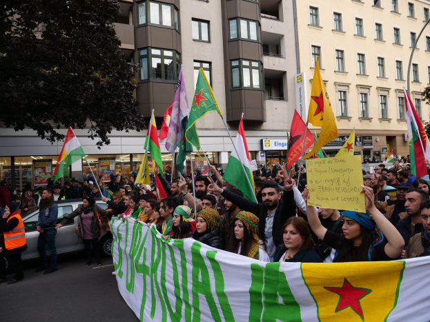 Eine große Gruppe von Menschen marschiert mit Flaggen und Spruchbändern die Straße entlang, mit einem geparkten Auto auf der rechten Seite und einem Baum auf der linken Seite, vor einem Hintergrund von Gebäuden mit Fenstern und Namensschildern, was auf eine algerische Demonstration hinweist.