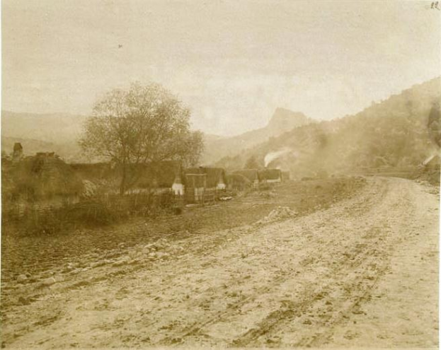 Ein altes Schwarz-Weiß-Foto einer Schotterstraße, die sich durch eine ländliche Landschaft schlängelt mit Häusern, Bäumen, Bergen und einem klaren Himmel.