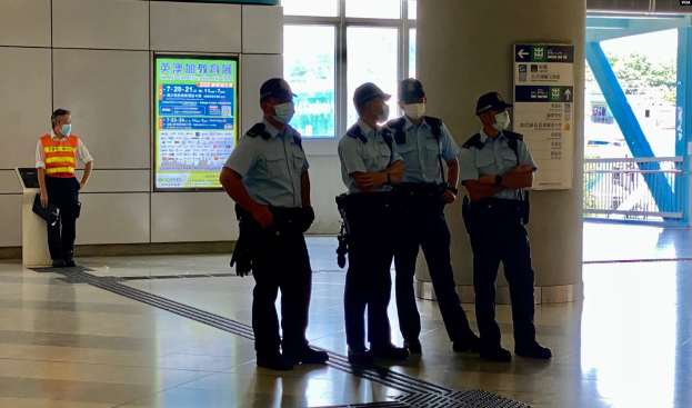 Eine Gruppe maskierter Polizisten steht vor einem Gebäude mit Glastüren, Schildern mit Text, einer Säule und einem Geländer, wobei durch die Glastüren Bäume und Fahrzeuge zu sehen sind.