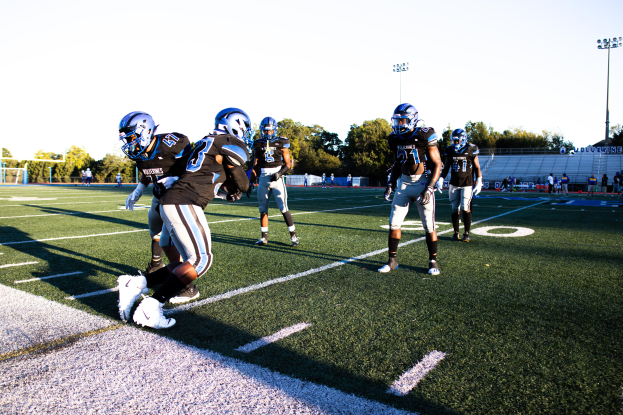 Eine Gruppe von Menschen, die Fußball auf einem von Bäumen, Laternen und einem klaren blauen Himmel umgebenen Feld spielen, wobei einige Spieler Helme und Schuhe tragen.