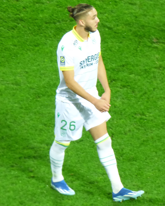 Ein Fußballspieler steht auf einem saftig grünen Feld und trägt ein weißes T-Shirt, Shorts, Socken und Turnschuhe.