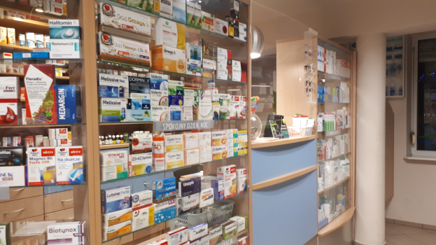 Pharmacy shelves stocked with various medicine bottles, boxes, and containers, featuring a central pillar and a glass window on the right.