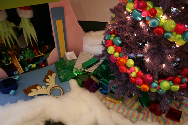 Eine Indoor-Szene mit einem Weihnachtsbaum mit Lichterketten und Dekorationen auf der rechten Seite, Geschenkboxen und Dekorationsgegenstände in der Mitte und einer Hütte mit Dekorationen und Watte in der oberen linken Ecke, mit einer Wand im Hintergrund und Watte und Wolle Stoff unten.