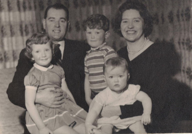 Eine Familie mit fünf Personen, darunter ein Mann in Anzug und Krawatte auf der linken Seite, eine Frau auf der rechten Seite und drei strahlende Kinder, die gemeinsam posieren.