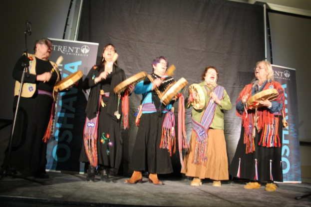 Eine Gruppe von Frauen in bunten Kleidern, die singen und Trommeln halten, vor einem schwarzen Vorhang und zwei blauen Rollbannern.