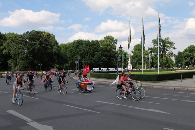 Eine Gruppe von Menschen, die auf Fahrrädern eine von Bäumen gesäumte Straße entlangfahren, während der Pariser Radtour 2016. Im Hintergrund sind Laternenpfähle, Flaggenpfähle, bewölkter Himmel und Fahrzeuge zu sehen.