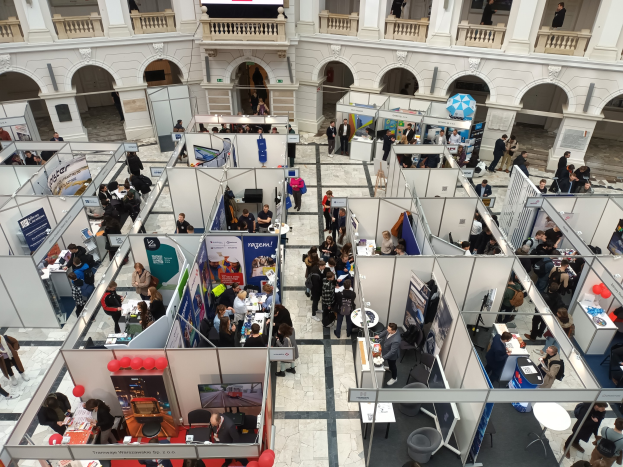 Große Gruppe von Teilnehmern auf einer internationalen Fachmesse mit Ständen, Bannern, Tischen, Stühlen und Ballons in einem Raum mit architektonischen Bögen, Säulen, Geländern und einem Bildschirm im Hintergrund.