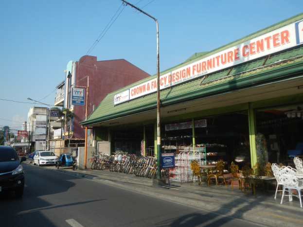 Eine belebte Stadtstraße mit fahrenden Fahrzeugen, parkenden Fahrrädern, Gebäuden mit Schildern, Laternenmasten, Strommasten mit Drähten, einem sichtbaren Himmel im Hintergrund und einem Möbelgeschäft im Vordergrund.