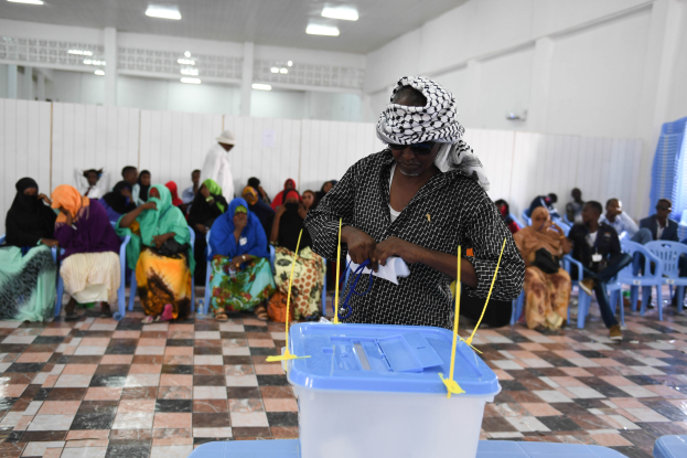 Eine Frau gibt ihre Stimme an einer Wahlurne in Mogadischu, Somalia ab, stehend an einem Tisch mit einer Box, umgeben von sitzenden Menschen und einer stehenden Person im Hintergrund, mit Deckenlampen, die den Raum beleuchten.