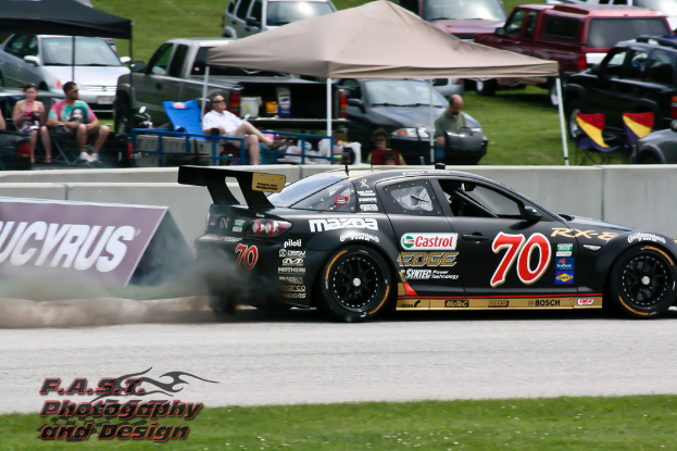 Ein schwarzer Rennwagen auf einer Straße mit Gras unten, eine Tafel mit Text links, Menschen unter Zelten sitzend und andere Autos im Hintergrund.