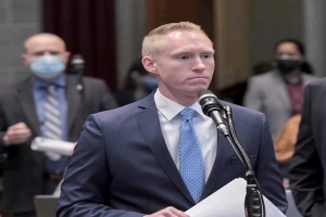 Ein Mann in Anzug und Krawatte spricht an einem Rednerpult mit Papieren, während andere hinter ihm stehen und eine Vorhangwand im Hintergrund zu sehen ist, in Bezug auf Vergewaltigungsvorwürfe.