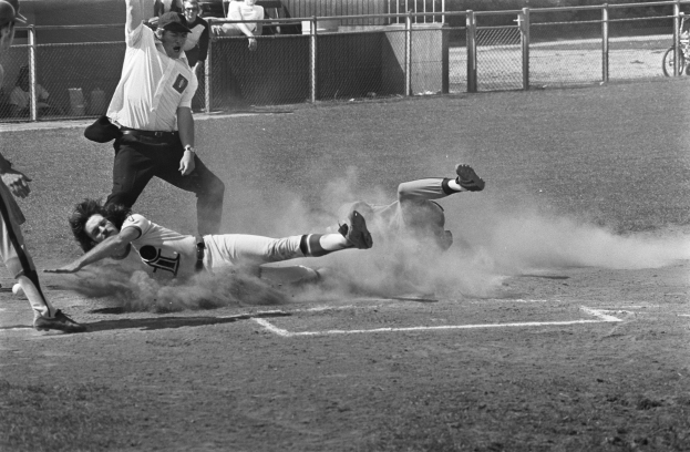 Ein Schwarz-Weiß-Bild eines Baseball-Spielers, der auf Home Plate rutscht, umgeben von einer Gruppe von Menschen auf dem Feld, mit einem Zaun, einem Fahrrad, Zuschauern, einem Gebäude, Bäumen und einem bewölkten Himmel im Hintergrund.
