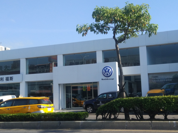 Volkswagen-Händler mit Glaswänden, parkende Autos davor, umgeben von Pflanzen und einem Baum, unter einem sichtbaren Himmel.
