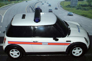 Weißer Polizeiwagen vor einer belebten Straße mit mehreren Fahrzeugen, Gras und Bäumen auf beiden Seiten, aufgenommen außerhalb einer Stadt.