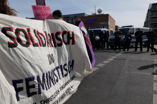 Gruppe von Menschen marschiert auf einer Straße und hält ein "Solidarität und Feminismus"-Schild, mit parkenden Fahrzeugen, Gebäuden, einer Parabolantenne und einem klaren blauen Himmel im Hintergrund.