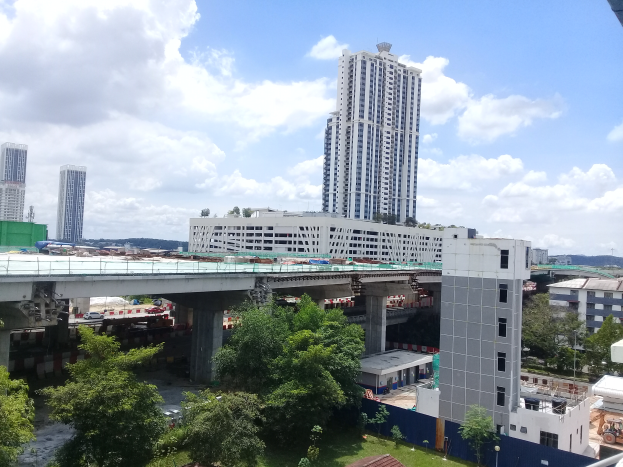 Eine Stadtansicht von einem Gebäude aus, mit Gebäuden, Bäumen, Gras, Fahrzeugen, einer Brücke, Pfählen und einem bewölkten Himmel, mit einer Baustelle für einen neuen Wohnkomplex im Vordergrund.
