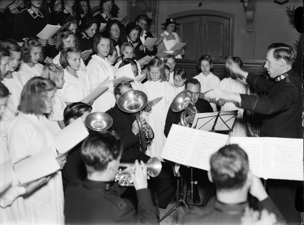 Eine Gruppe von Menschen vor einem Chor stehend, jeder hält ein Buch und spielt verschiedene Musikinstrumente, dargestellt in Schwarz-Weiß.